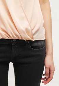 Léková halenka v broskvové barvě s detailem na staženém pase, zkombinovaná s černými džíny. Hladká textura a volný střih, se zapínáním na zavazování vpředu.