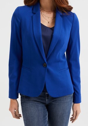 Kvinde iført en tætsiddende royalblå blazer over en mørkeblå top, kombineret med blå jeans og lagdelte guldkæder.