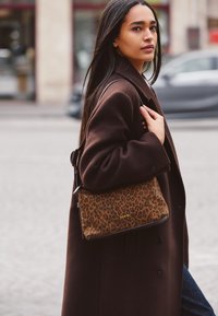 Brown oversized coat over a black top, carrying a leopard print bag with a smooth texture and dark accents, in an outdoor urban setting.