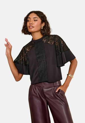 Schwarze Bluse mit Spitzenverzierungen, kurzen Capeärmeln und einem zentralen strukturierten Panel, kombiniert mit burgunderfarbenen Lederhosen. Die Accessoires umfassen ein goldenes Armband.