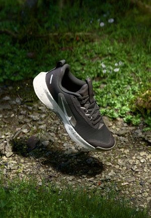 Sort-hvid Nike løbesko ophængt over klippefyldt terræn med græs og små blomster i baggrunden.