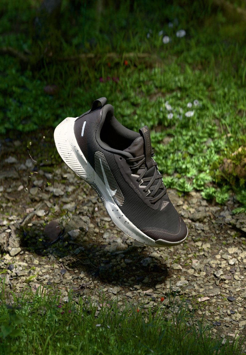 Chaussure de course Nike noire et blanche suspendue au-dessus d'un terrain rocheux avec de l'herbe et de petites fleurs en arrière-plan.