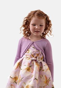 Cardigan viola con bordo in pizzo sopra un vestito floreale caratterizzato da grandi fiori pesca e bianchi su uno sfondo viola chiaro.