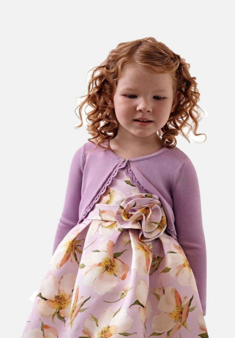 Cardigan viola con bordo in pizzo sopra un vestito floreale caratterizzato da grandi fiori pesca e bianchi su uno sfondo viola chiaro.