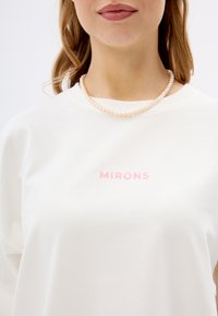 Witte katoenen sweatshirt met ronde hals, met roze geborduurde tekst "MIRONS" op de borst en gedragen met een parelketting.