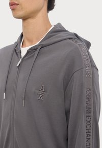 Šedá mikina na zips s mäkkou textúrou, so klokankou, kapucňou na šnúrku a vyšitými logami na hrudi a rukávoch.