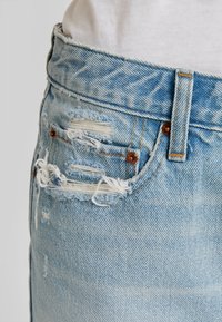 Jeans in denim azzurro chiaro con dettagli consumati, molteplici strappi, bordi sfilacciati e cuciture marroni a contrasto. Vestibilità a vita media.