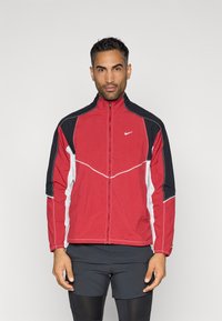 Sarkans un melns zip-up jaciņš ar baltiem akcentiem, kuram ir augsts apkakle un kontrastējošas šuves. Materiāls izskatās viegls un elpojošs.