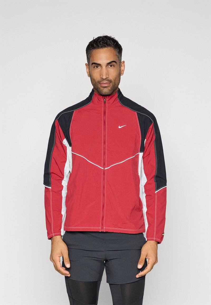 Sarkans un melns zip-up jaciņš ar baltiem akcentiem, kuram ir augsts apkakle un kontrastējošas šuves. Materiāls izskatās viegls un elpojošs.