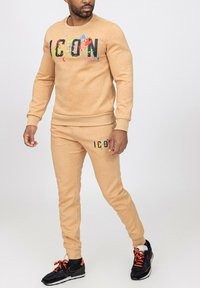 Sweatshirt en coton beige avec le logo "ICON" en impression éclaboussure colorée, associé à un jogger assorti avec détails du logo. Porté avec des baskets noires.