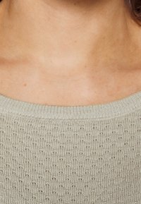 Pull en maille vert clair avec un large décolleté, présentant un motif texturé et un ourlet côtelé. Tissu doux avec un léger éclat.