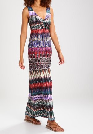 Femme portant une robe maxi sans manches à col en V avec des motifs géométriques et rayures multicolores, associée à des sandales plates marron.