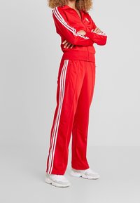 Röd atletisk träningsoverall med vita tre-randsaccenter på armar och sidor, zip-up jacka och löst sittande byxor av slät tyg.