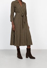 Abito a scacchi marrone e nero con scollatura a V, vita annodata e maniche lunghe. Abbinato a stivali col tacco marroni. Tessuto con finitura liscia.