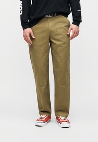 Άτομο που φοράει παντελόνι ελιάς με ενισχυμένες επιχρίσεις στα γόνατα, μαύρο πουκάμισο με μακριά μανίκια και κόκκινα αθλητικά παπούτσια χαμηλού τύπου σε απλό λευκό φόντο.