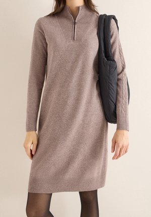 Kobieta w długiej sukience z dzianiny w kolorze wrzosowym z półgolfem na zamek oraz czarną pikowaną kamizelką przewieszoną przez ramię.