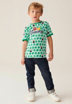 T-shirt verde a maniche corte con una grafica "Super Lucky" e pattern di trifogli e tucani sparsi, abbinato a jeans scuri e scarpe da ginnastica bianche.