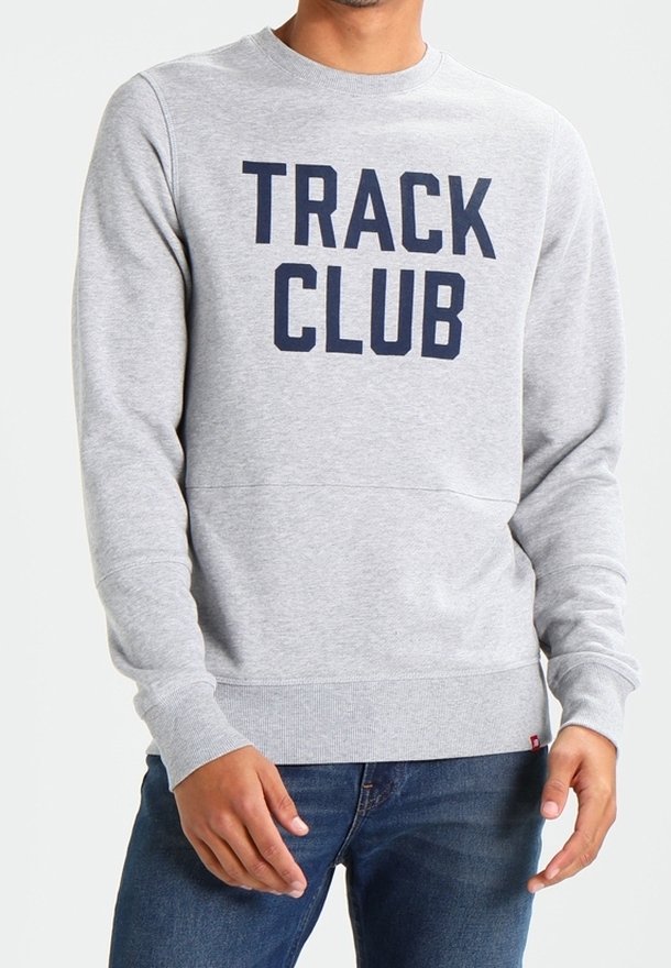 Homme portant un sweat-shirt gris clair avec l'inscription "TRACK CLUB" en grandes lettres bleu marine, assorti à un jean bleu.