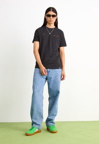 Černé tričko s krátkým rukávem s logem Puma, světle modré široké džíny a zelené nízké tenisky s růžovými akcenty. Model nosí sluneční brýle.