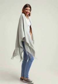 Grå sjal med fransade kanter, bärs över en vit topp och blå jeans, på en neutral bakgrund. Mjuk tyg, avslappnad hängning och lagom design.