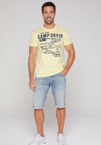 Camiseta amarilla clara con texto impreso en negro, combinada con pantalones cortos de mezclilla azul claros y zapatillas blancas. Posición plana contra un fondo sencillo.