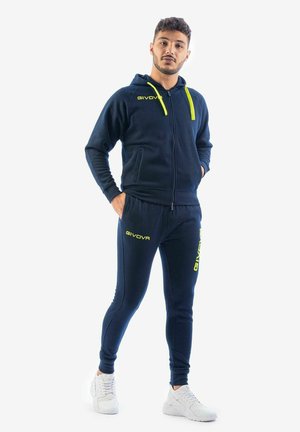 Uomo in piedi con le mani in tasca che indossa una tuta Givova blu navy con accenti gialli e scarpe da ginnastica bianche su sfondo bianco.