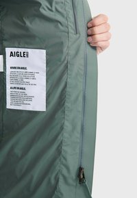 Veste verte légère avec une texture lisse. Dispose d'une fermeture éclair sur le côté et d'une étiquette blanche avec du texte noir à l'intérieur.
