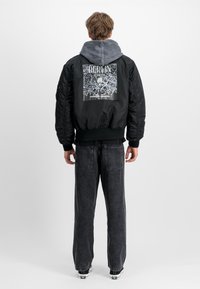 Chaqueta bomber negra con capucha gris, con un gráfico blanco de un mapa de Berlín en la parte de atrás. Usada sobre pantalones de mezclilla oscuros, de corte casual.