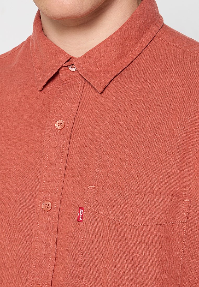 Chemise ajustée couleur rouille faite de tissu texturé. Présente un col pointu, deux boutons sur la patte de boutonnage et une poche poitrine avec une étiquette de logo.