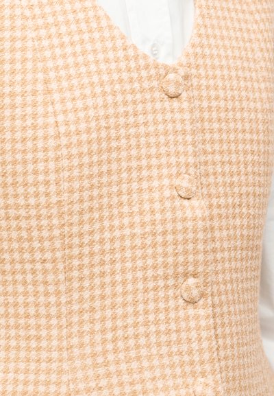 Μπεζ γιλέκο με λευκό μοτίβο houndstooth και κουμπιά καλυμμένα με ύφασμα, φορεμένο πάνω από ένα λευκό πουκάμισο με γιακά.
