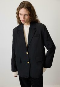 Melnā žakete ar pielāgotu piegriezumu, zelta pogām, divām priekšējām kabatām, uzvilkta virs krēmkrāsas kabelī aušanas džempera. Gluda tekstūra, strukturēts dizains.