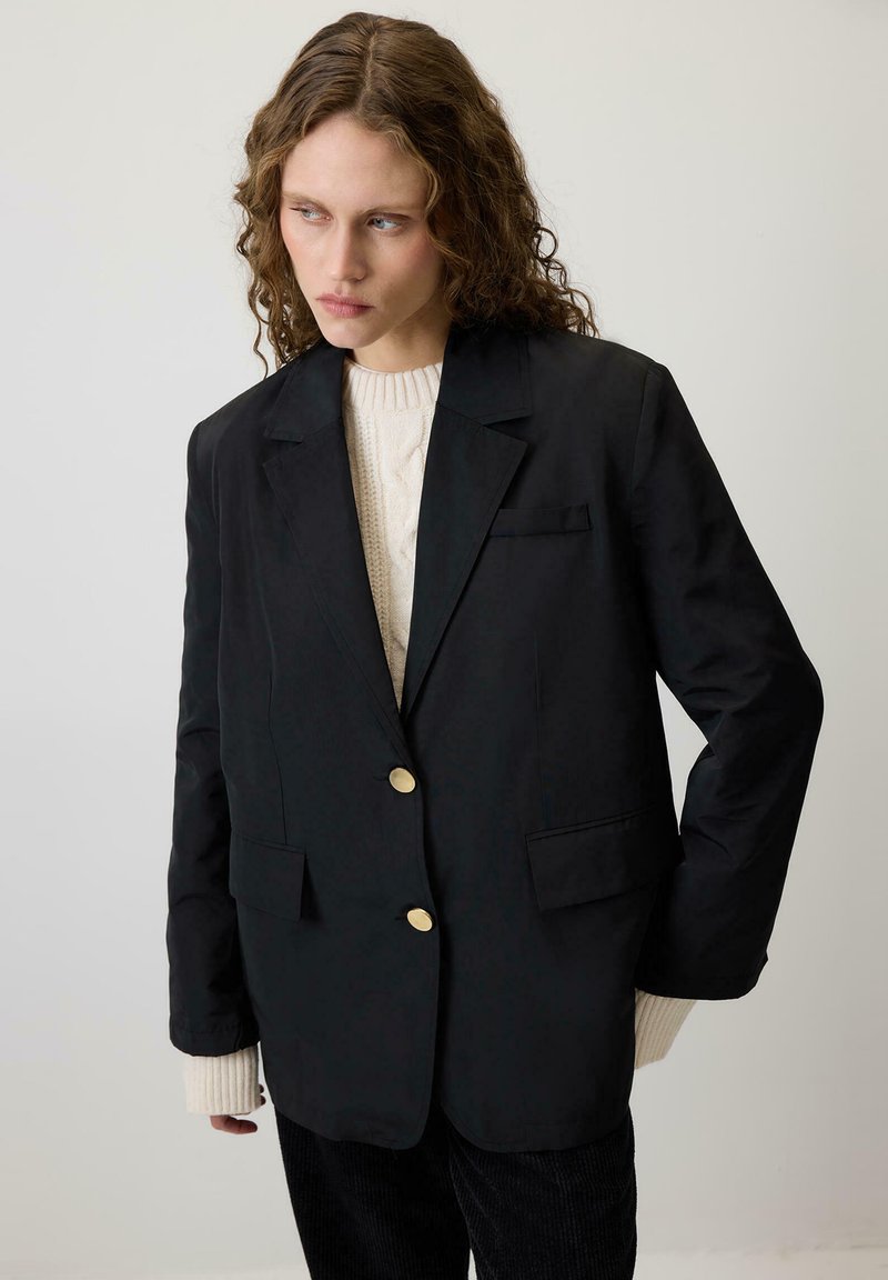 Melnā žakete ar pielāgotu piegriezumu, zelta pogām, divām priekšējām kabatām, uzvilkta virs krēmkrāsas kabelī aušanas džempera. Gluda tekstūra, strukturēts dizains.