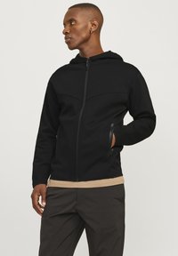 Svart hettejakke med glidelås, med en jevn tekstur, en tettsittende design og sidelommer. Brukt over en beige topp.