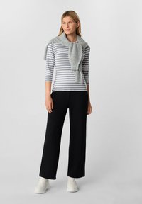 Lange schwarze Hosen kombiniert mit einem grau gestreiften Langarmshirt und einem grauen Strickschal. Das Model trägt weiße Turnschuhe und steht vor einem neutralen Hintergrund.