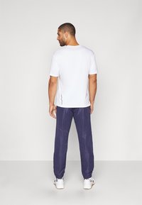 T-shirt blanc à manches courtes et pantalon de sport bleu marine avec des poignets élastiques, porté avec des baskets blanches. Design simple, le matériau semble léger.