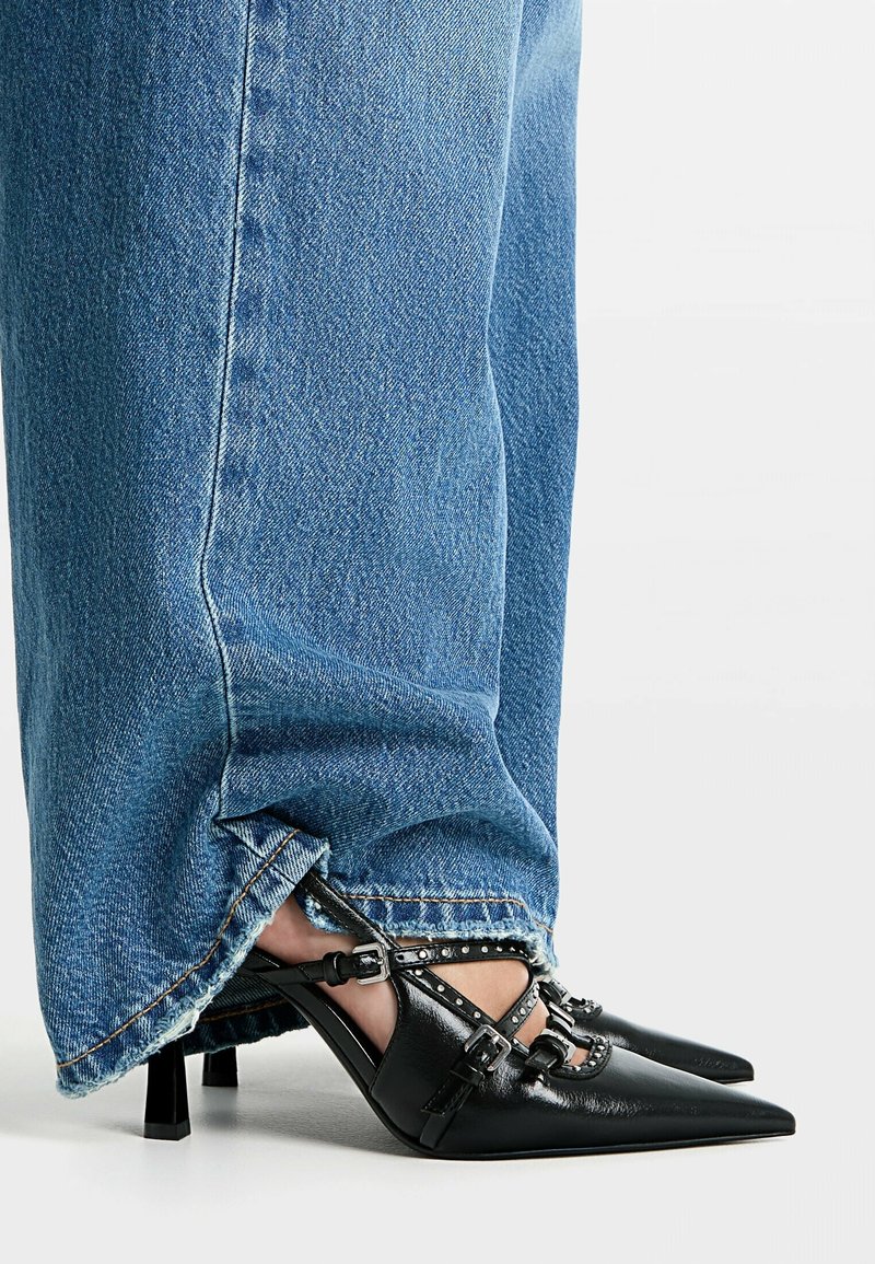 Chaussures à talons noirs à bout pointu avec brides autour de la cheville et clous argentés, associées à un jean en denim bleu à ourlet effiloché. Texture en cuir lisse et talon aiguille.
