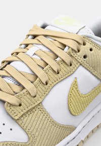 Tenisky Nike s bielou koženou základňou, žltými kordurovými akcentmi, textúrovanými panelmi a béžovými šnúrkami. Diskrétne logo na boku.