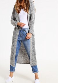 Lång gråstickad cardigan med öppen front, texturerad tyg och ribbade ärmar, bärs över en vit topp och blåuppvikta jeans.