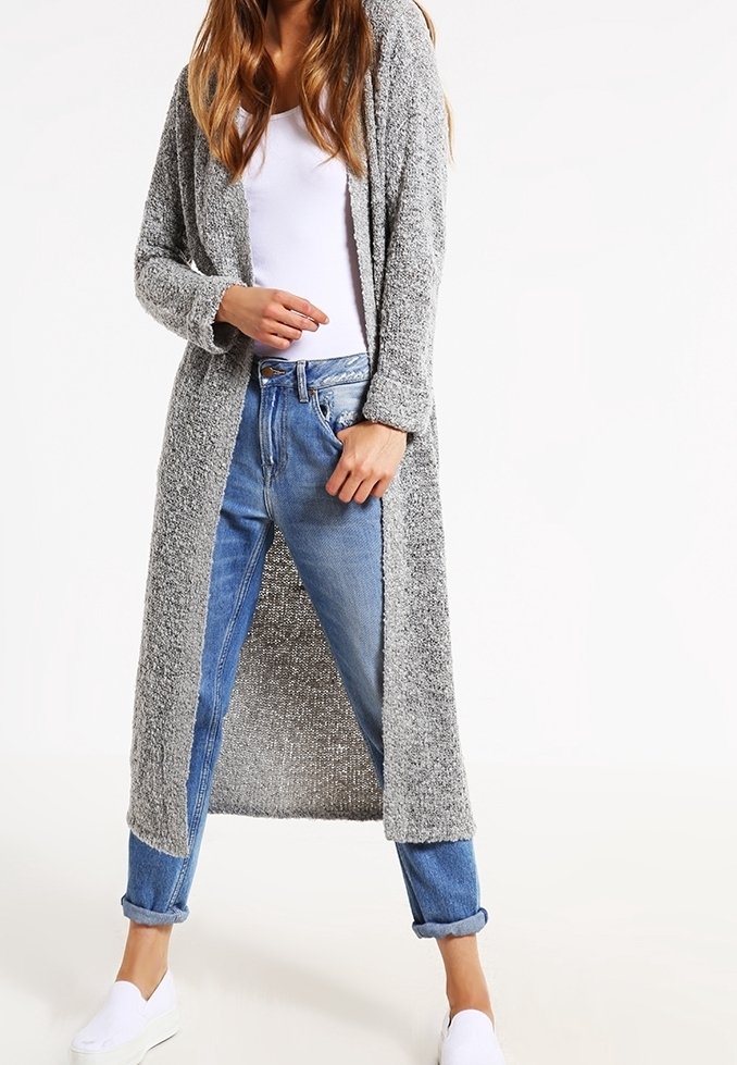 Lång gråstickad cardigan med öppen front, texturerad tyg och ribbade ärmar, bärs över en vit topp och blåuppvikta jeans.