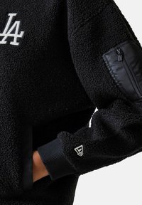 Schwarzer Fleece-Pullover mit einem weißen LA-Logo, einer Reißverschlusstasche am Ärmel und gerippten Bündchen für einen sicheren Sitz.