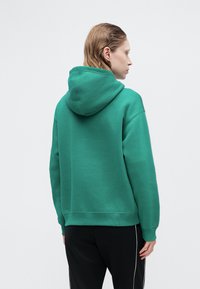 Zielona bluza z kapturem, materiał bawełniany, luźny krój, ściągacze w mankietach i na dole, minimalne detale przeszycia, noszona z ciemnymi spodniami.
