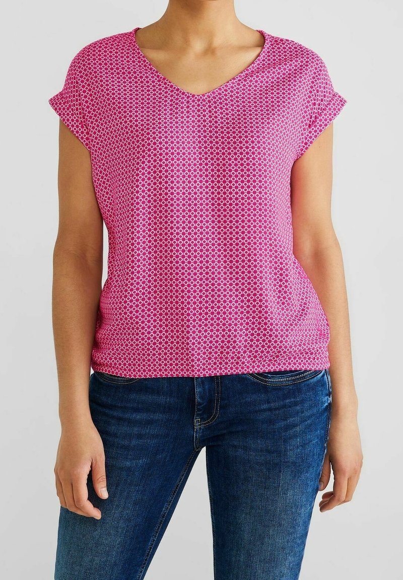 Femme portant un chemisier rose à manches courtes avec col en V à motifs et un jean bleu en denim, debout devant un fond clair uni.