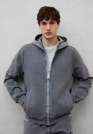 Joven con cabello rizado que lleva una sudadera gris con cremallera y pantalones a juego, de pie con las manos en los bolsillos contra un fondo liso.