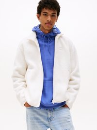 Witte fleec jas met hoge kraag en ritssluiting; gelaagd over een blauwe hoodie met een trekkoord. Lichtblauwe distressed jeans maken de outfit compleet.