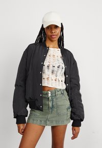 Nainen, jolla on pitkät letit, valkoinen lippalakki, musta bomber-takki, kerma-valkoinen neulecrop-top ja vihreä denim-minihelma yksinkertaista taustaa vasten.