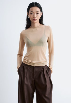 Jeune femme aux longs cheveux bruns portant un haut beige transparent à manches longues par-dessus un soutien-gorge, associé à un pantalon large marron foncé taille haute.