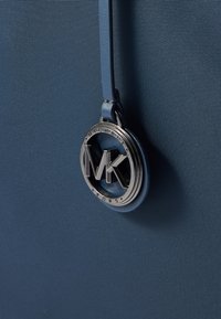 Niebieska teksturowana tkanina z okrągłym srebrnym zawieszką z logo Michael Kors, wiszącą na pasującym skórzanym pasku.