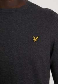 Tummanharmaa pusero teksturoidusta neuloksesta, jossa on pieni keltainen kotkalogo rinnassa pyöreän pääntien yläpuolella.