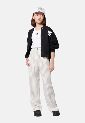 Zwarte varsityjack met wit logo, witte tanktop, gestreepte lichtbeige broek en zwarte instapschoenen. Witte pet met logo-accent.