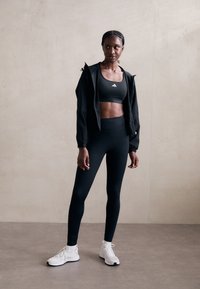 Sportiska sieviete, tērpusies melnā sporta krūšturī, melnās legingās, melnā jakā un baltās sporta apavos, stāv pret neitrālu sienu, skatoties sānis.